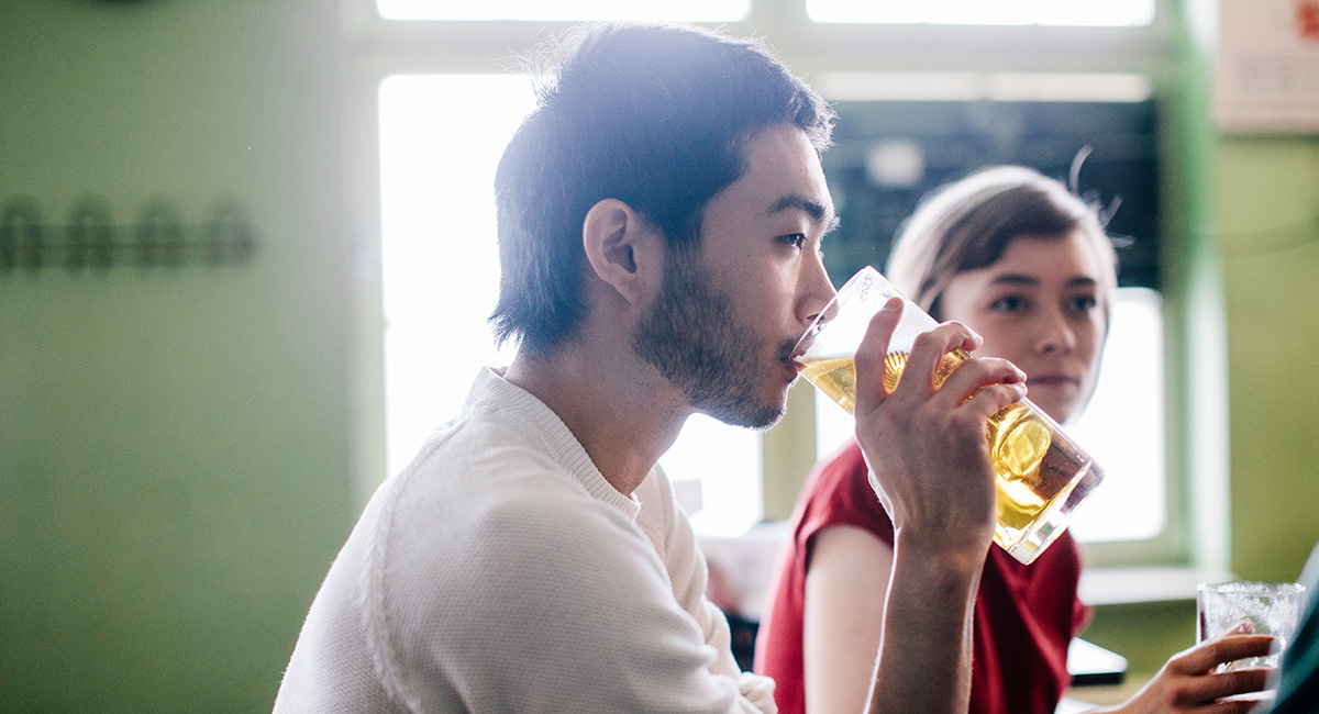 I förgrunden sitter en man och dricker ur ett glas, bakom honom sitter en kvinna i röd tröja.